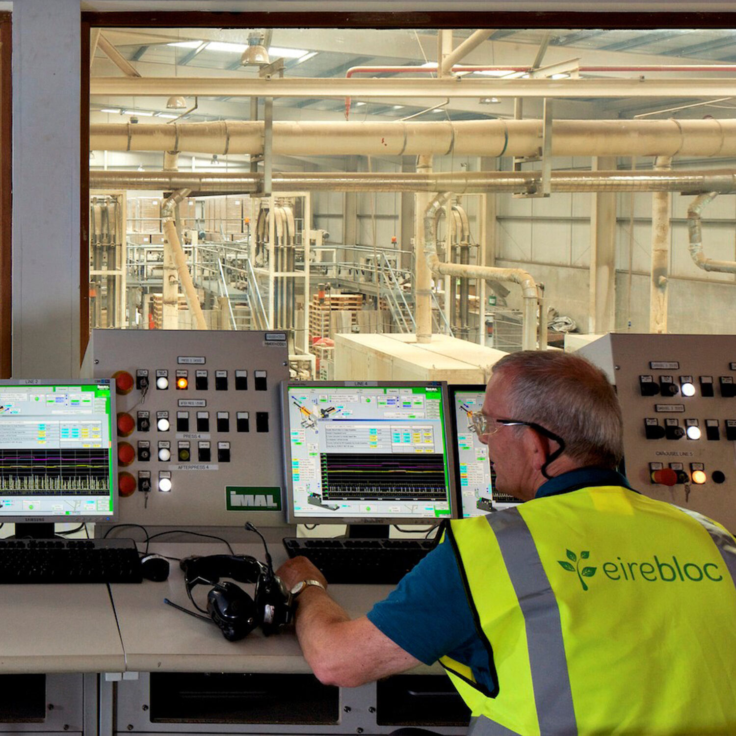 An Eirebloc operator in a hi-vis vest at the control room desk, with the production floor visible through the window.